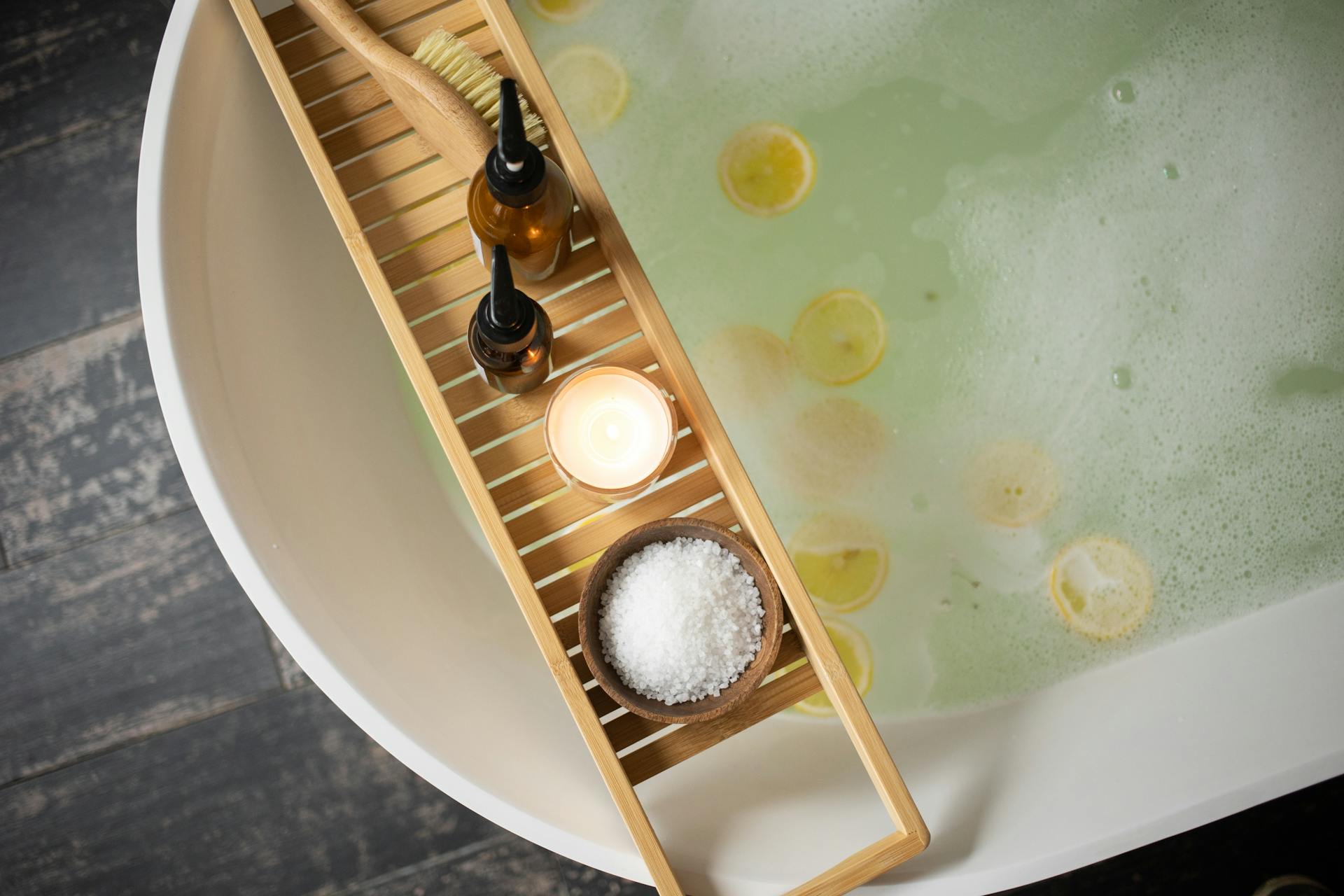 a photo of a full bath with lemon slices and a bath tray  with a lit candle, a bowl of bath salts, a scrubbing brush, and two bottles of hygiene items